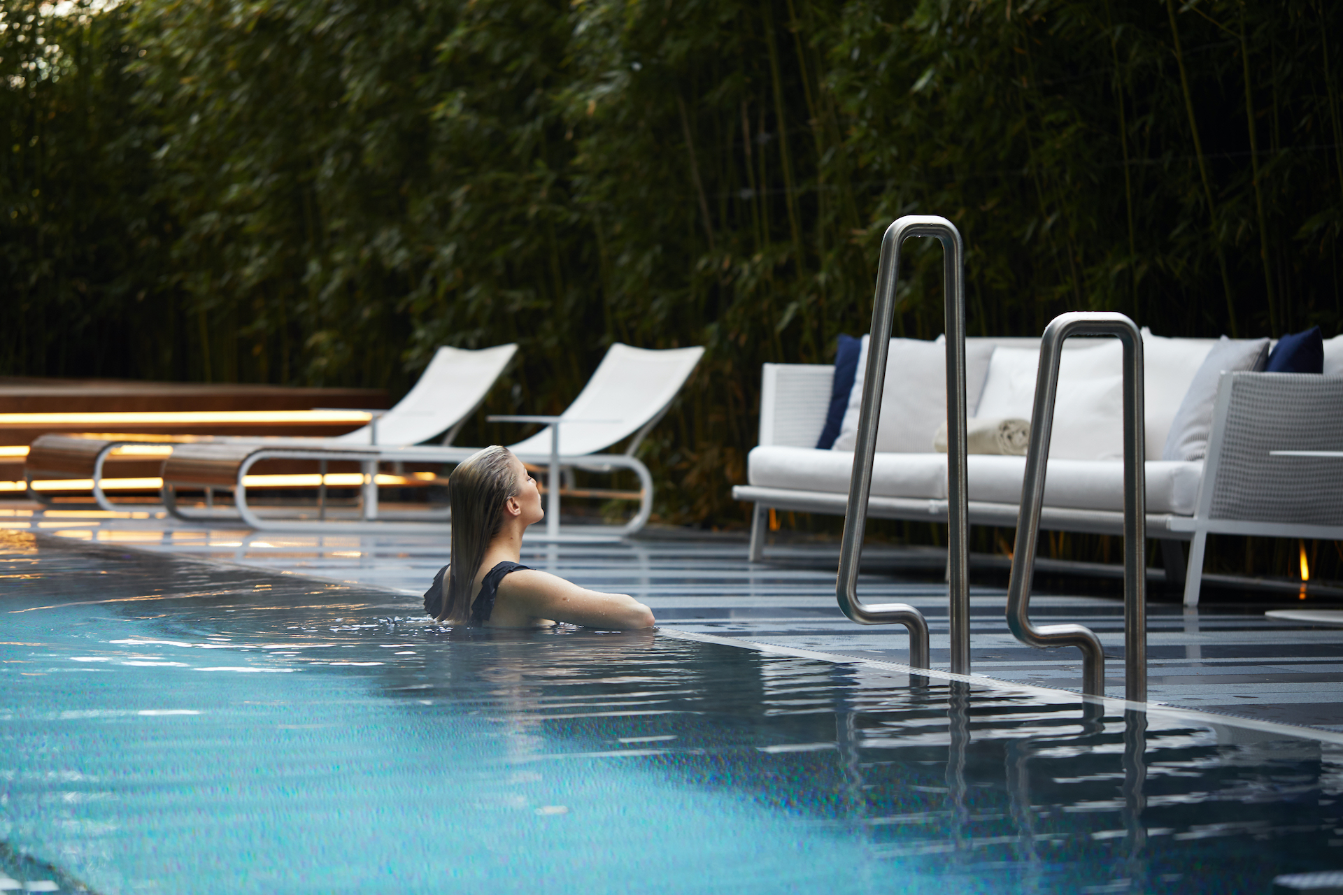 Tranquil poolside scene with a woman relaxing in water, surrounded by lush greenery and modern lounge furniture, epitomizing peaceful leisure in a luxury resort setting.