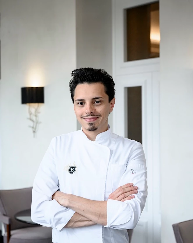 Confident chef in a crisp white uniform, posed in a warmly lit, elegant dining room, evoking professionalism and passion for culinary arts.