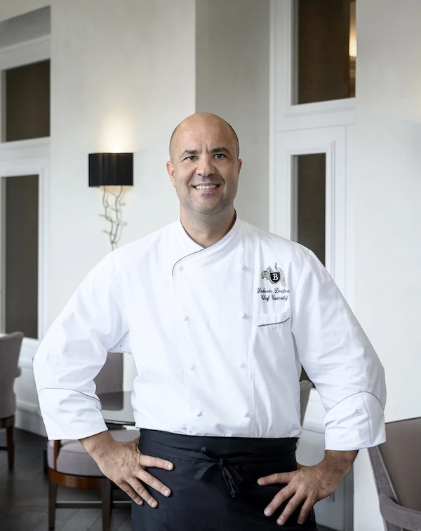 Confident chef in white jacket and black apron stands in an elegant dining room, offering a professional and welcoming atmosphere for culinary enthusiasts.