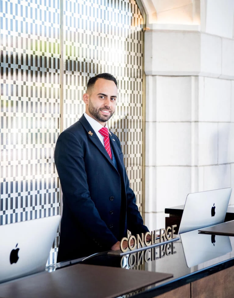 Professional hotel concierge in a blue suit at a sleek, modern desk with a mosaic tile backdrop, radiating a welcoming and attentive vibe for travelers.
